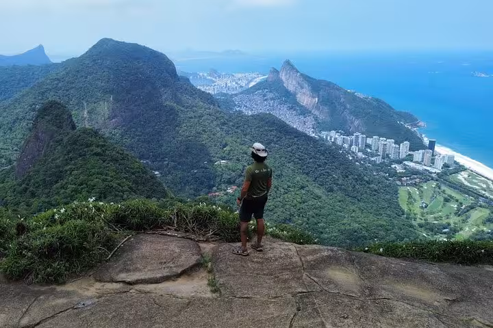 Hiker enjoys stunning panoramic views from a stone ledge in Tijuca Forest overlooking lush mountains and the ocean.