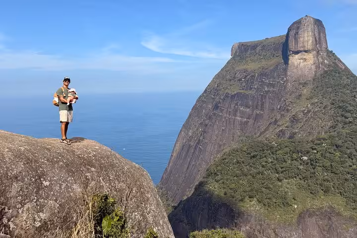 Hiker stands on a rock with panoramic views of Tijuca Forest and Pedra da Gávea under clear blue skies.