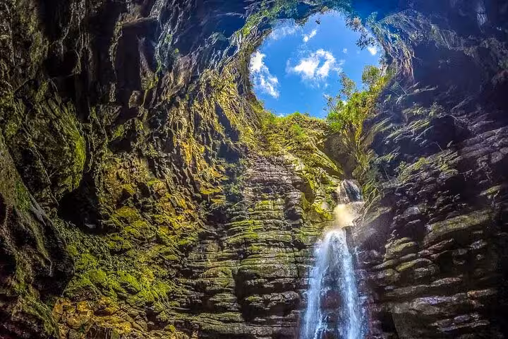 Explore the stunning Hole of the Father waterfall framed by lush greenery and rugged rock formations under a bright sky.