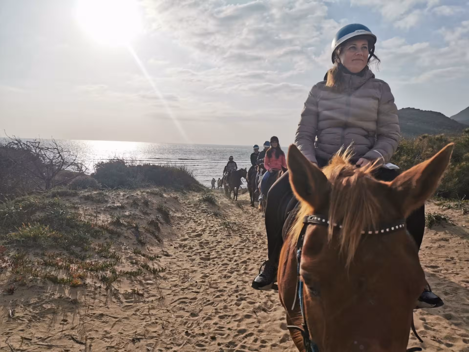 Riders enjoy a horseback tour near Lake Baratz in Alghero, with the sun setting over the shimmering sea.