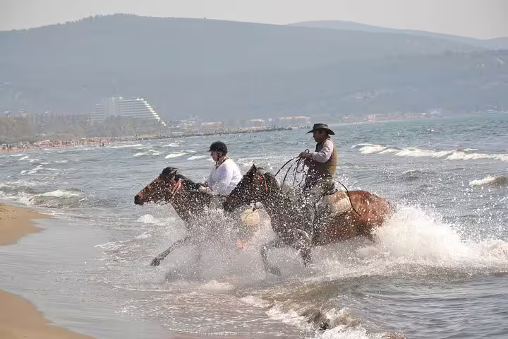 Two riders on horseback splashing through waves on a scenic beach near Kusadasi, perfect for adventurous cruise guests.