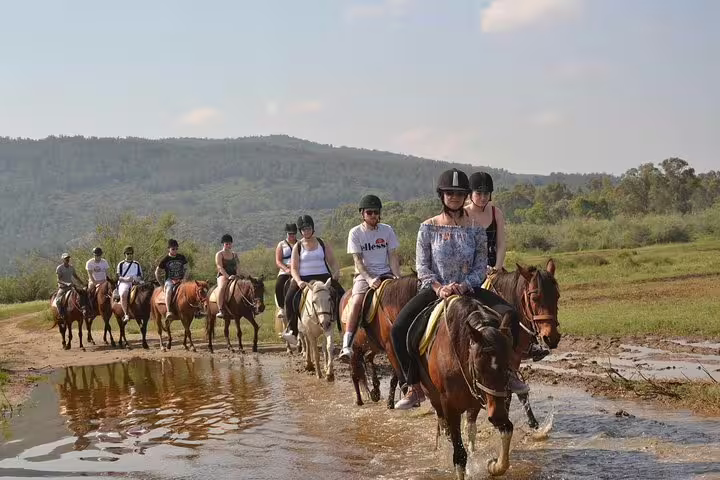 Riders on horseback cross a shallow stream with picturesque mountain views during a tour from Kusadasi Port.