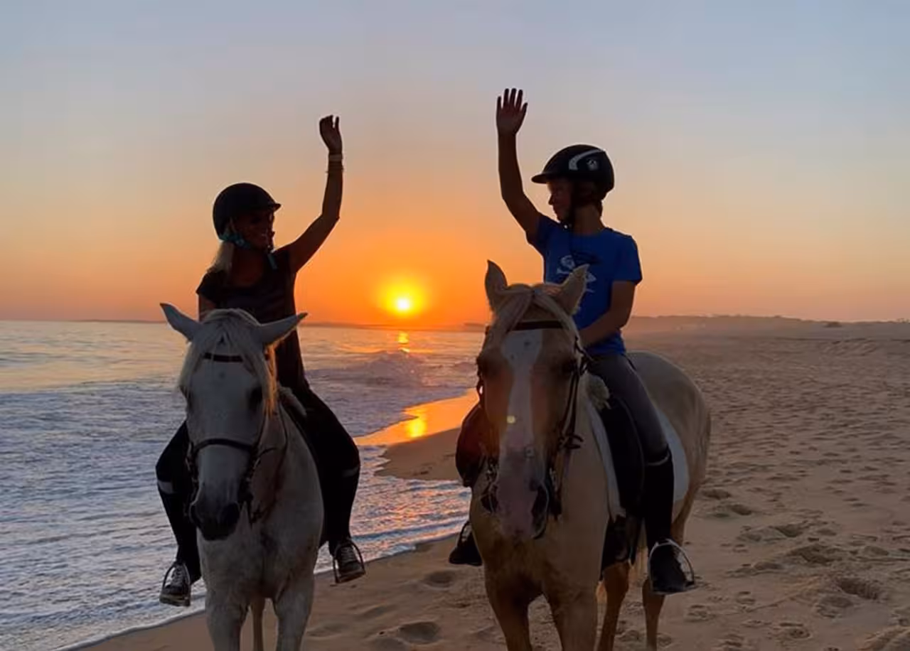 Two people on horseback waving against a stunning sunrise on Quarteira beach, perfect for an unforgettable ride.