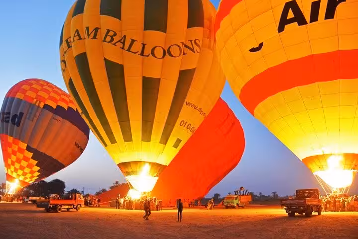 Hot air balloons glowing during pre-dawn launch in Luxor, Egypt, with hotel pickup and drop-off included
