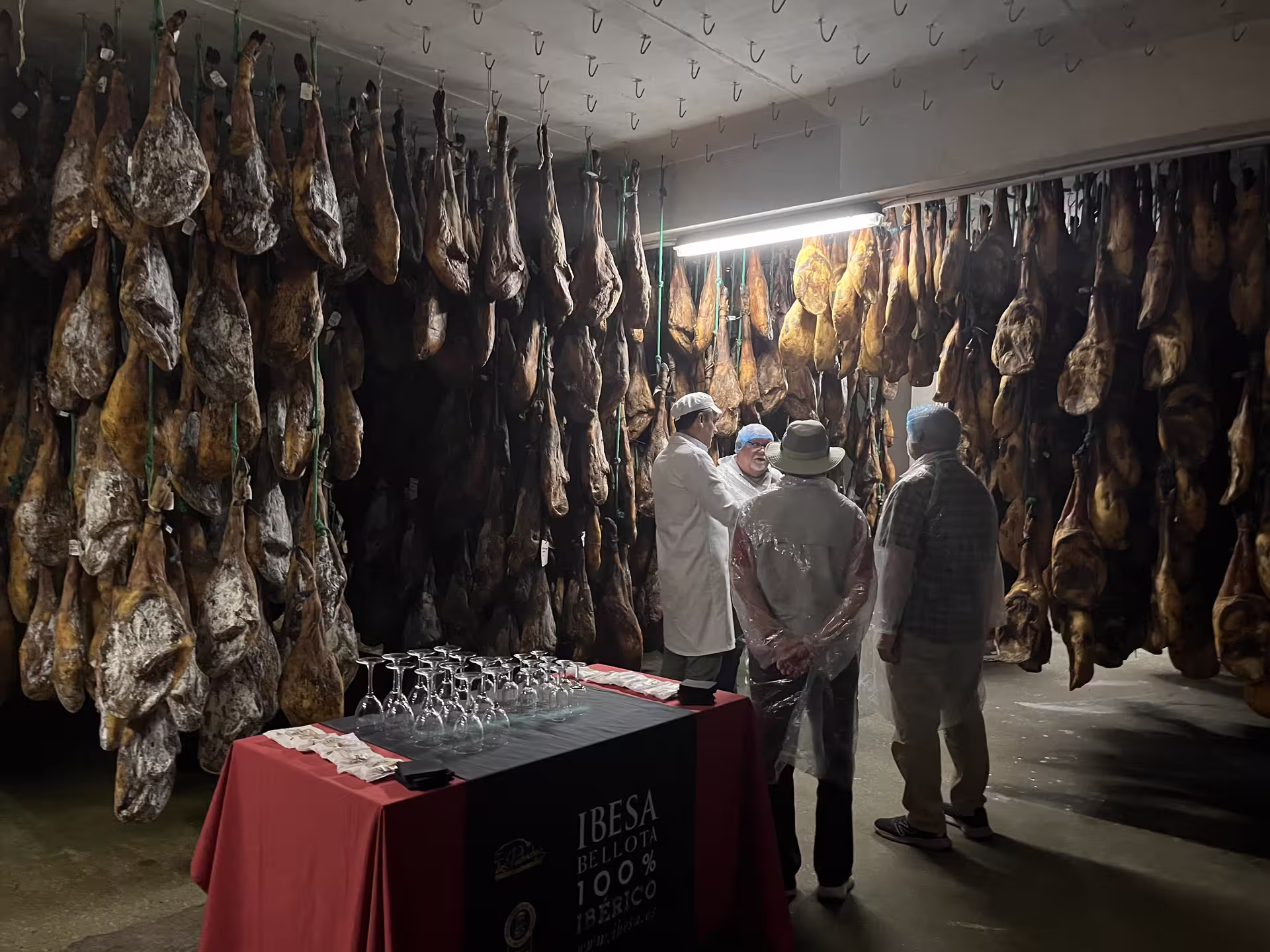 Group touring Iberian ham cellar with hanging cured meats, showcasing traditional Spanish delicacies.