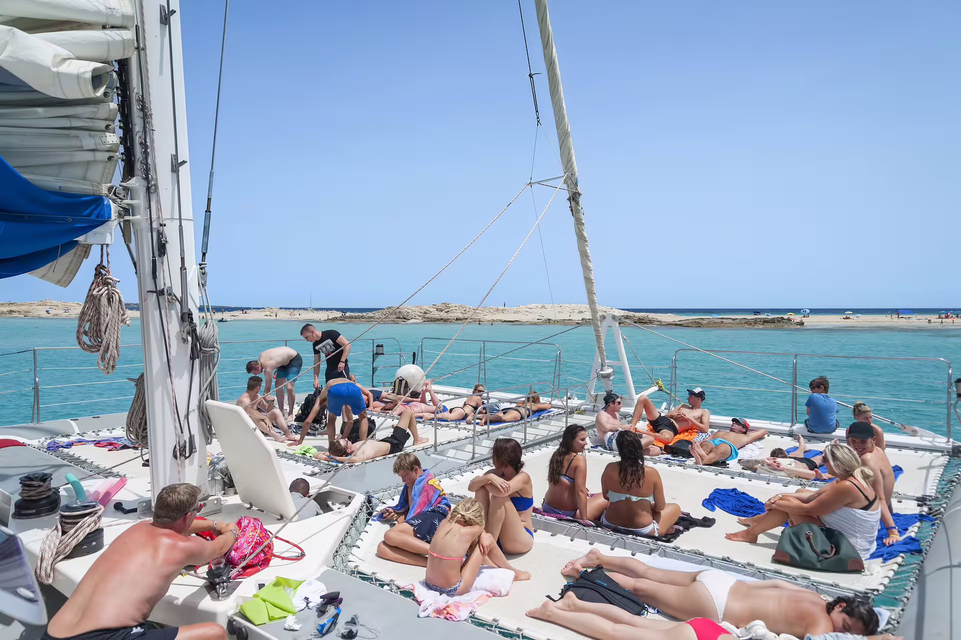 Guests sunbathing on Ibiza catamaran at Illetas, Formentera, with turquoise water and onboard lunch