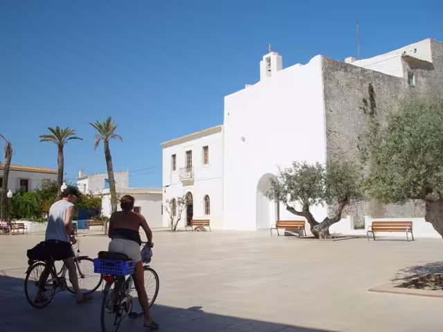 Cyclists crossing a sunny Ibiza village plaza near a white church on the Ibiza bus tour with fast ferry and guide