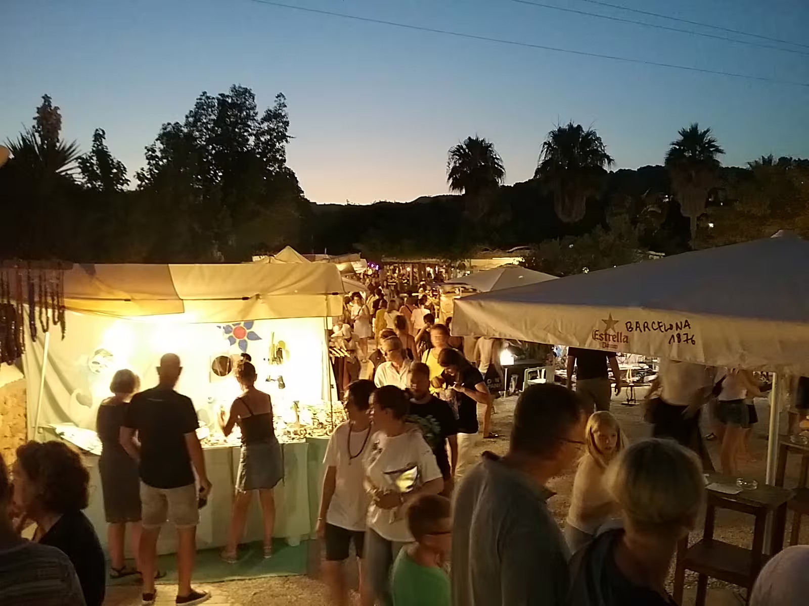 Crowds browsing Las Dalias hippie market at twilight on Ibiza sunset tour by bus, with lit stalls and palm trees