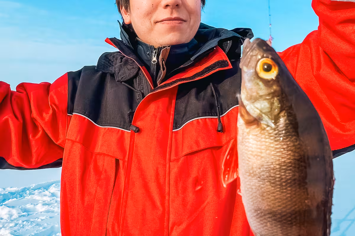 Proud ice angler in red gear displays freshly caught fish against a clear blue sky on frozen lake adventure.