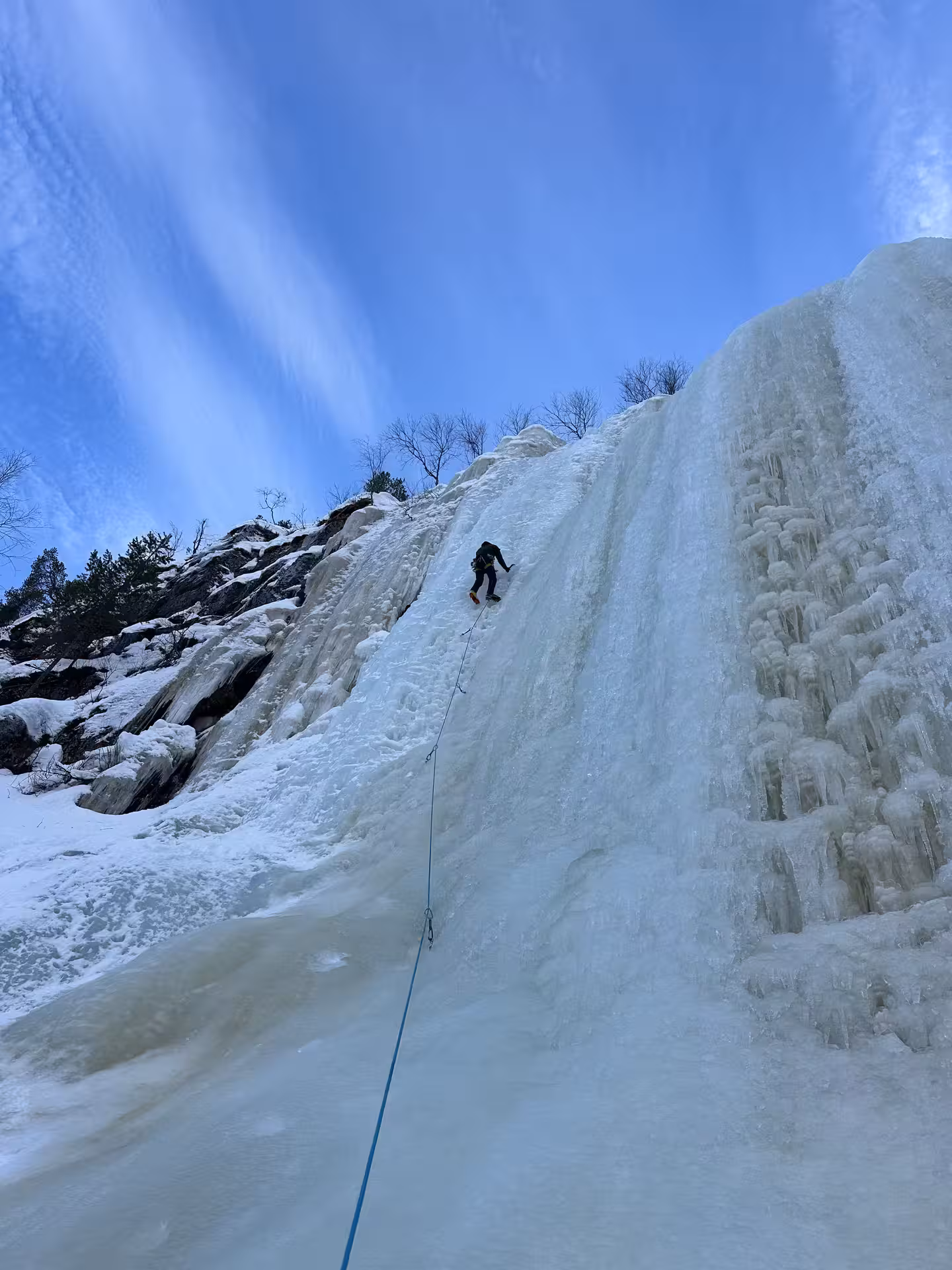 Solo ice climber ascending a steep frozen waterfall under a clear blue sky in Rovaniemi's winter landscape.