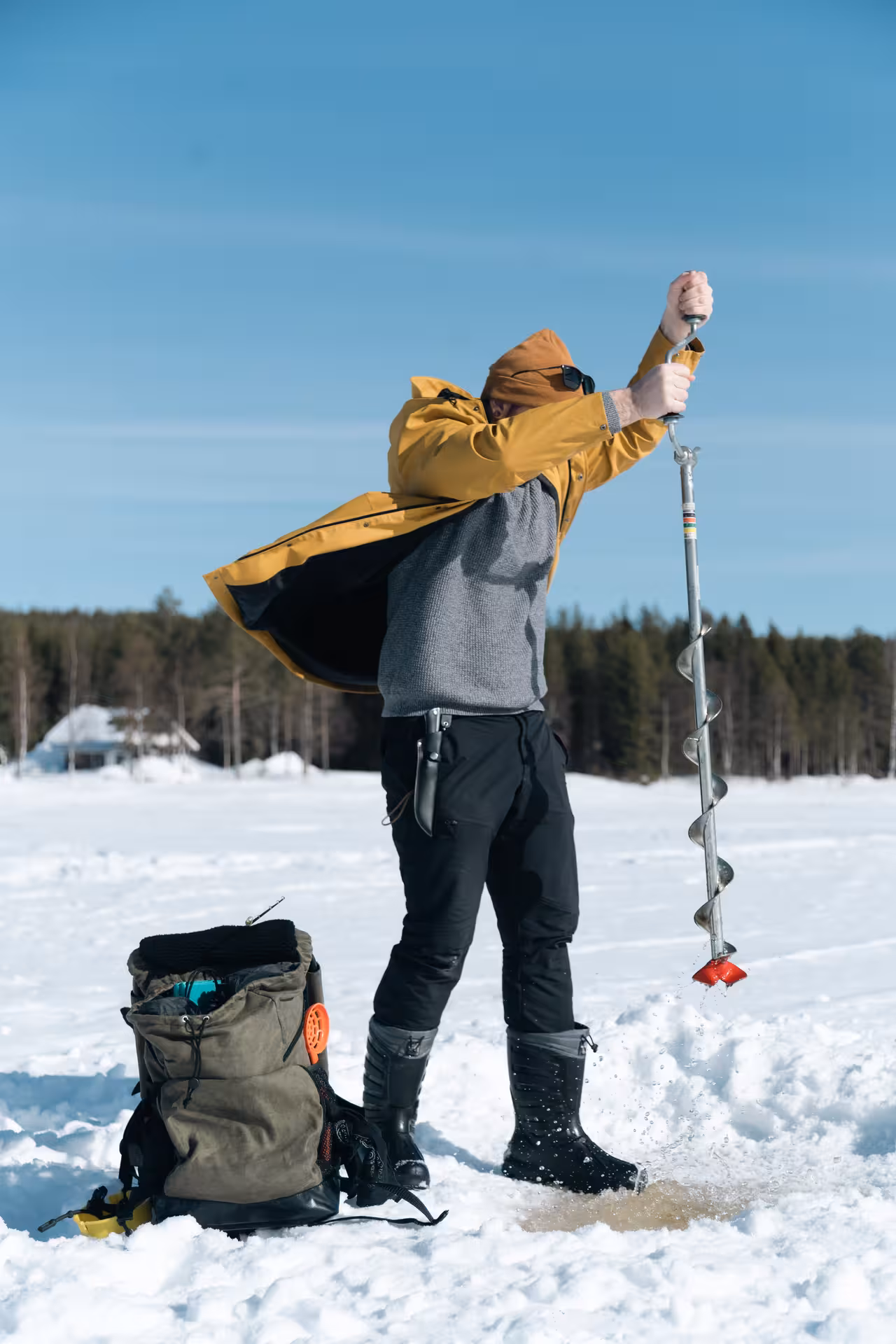 Ice fishing enthusiast drills into frozen lake with auger, preparing for a local fishing experience.
