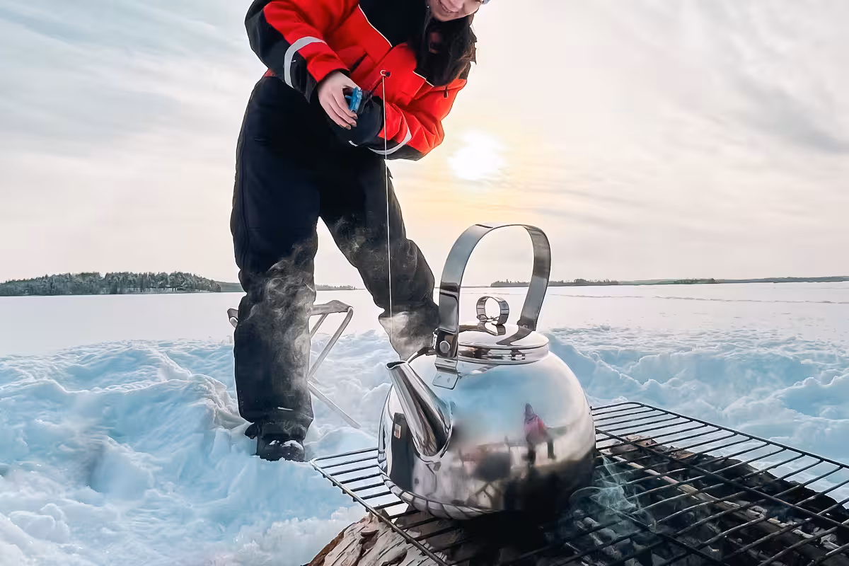 Local ice angler in red jacket enjoys tea break with kettle on grill during serene frozen lake fishing tour.