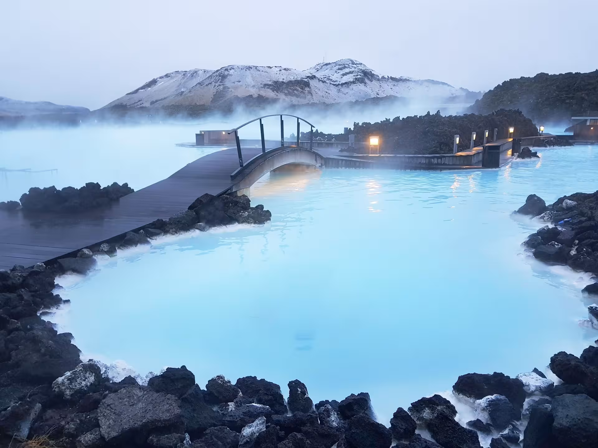 Relaxing Blue Lagoon geothermal spa with mist and snowy mountains on Iceland 6-Day Stopover Tour.