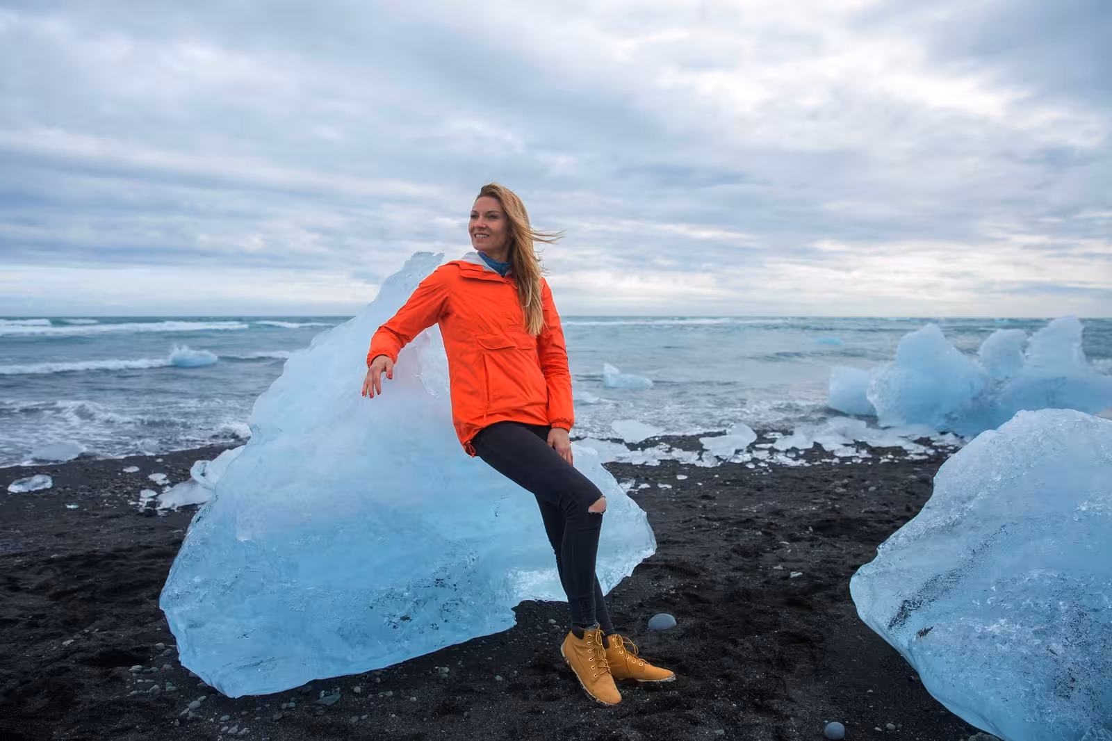 Woman in orange jacket sitting on ice at Diamond Beach, Iceland, during a self-drive tour.