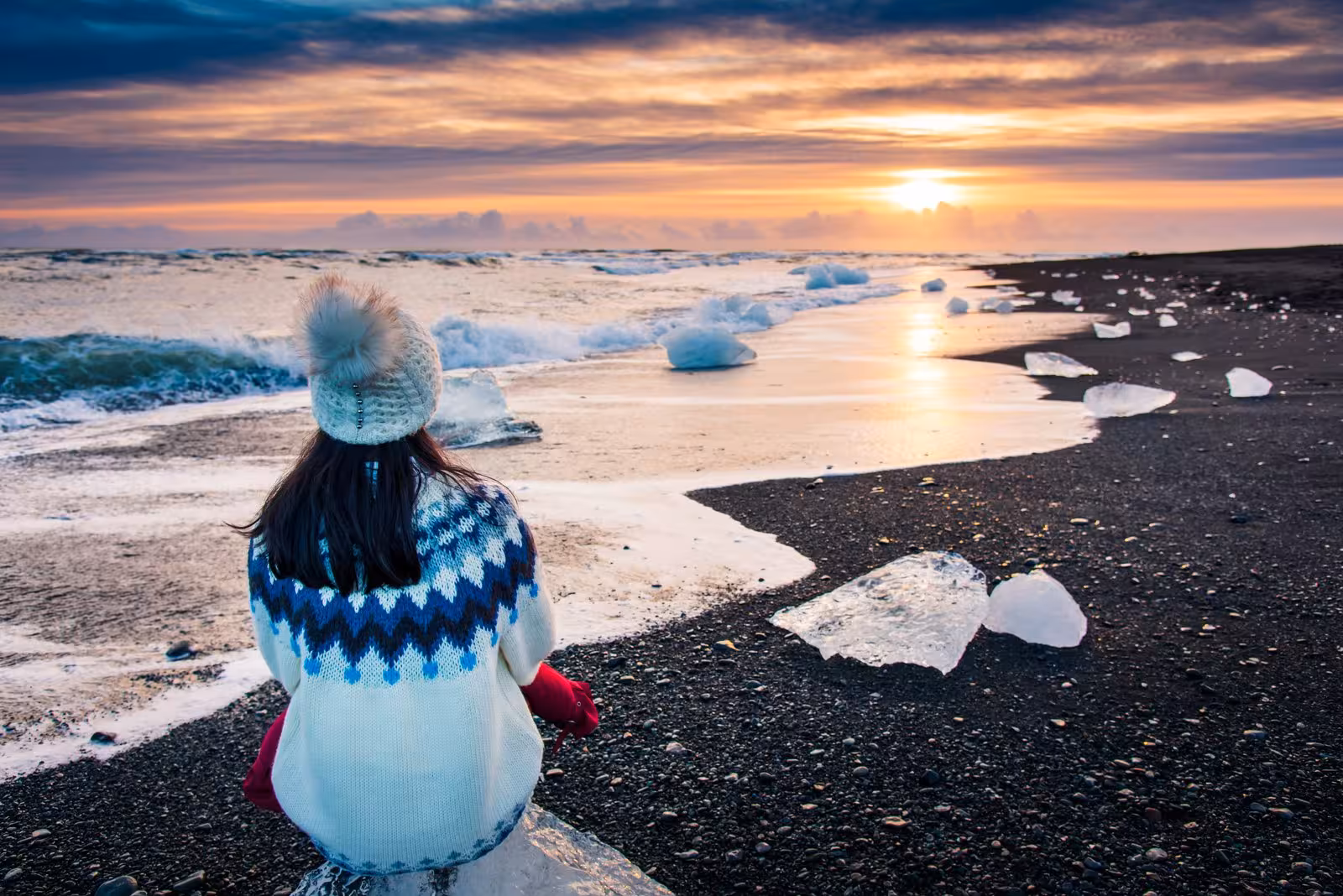 Person in cozy sweater watching sunset over Iceland's Diamond Beach with scattered icebergs on the Grand Tour.