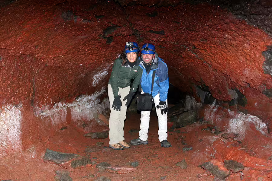 Two adventurers in helmets explore the vibrant red and rocky interior of Iceland's Leidarendi Lava Tunnel on a small group tour.