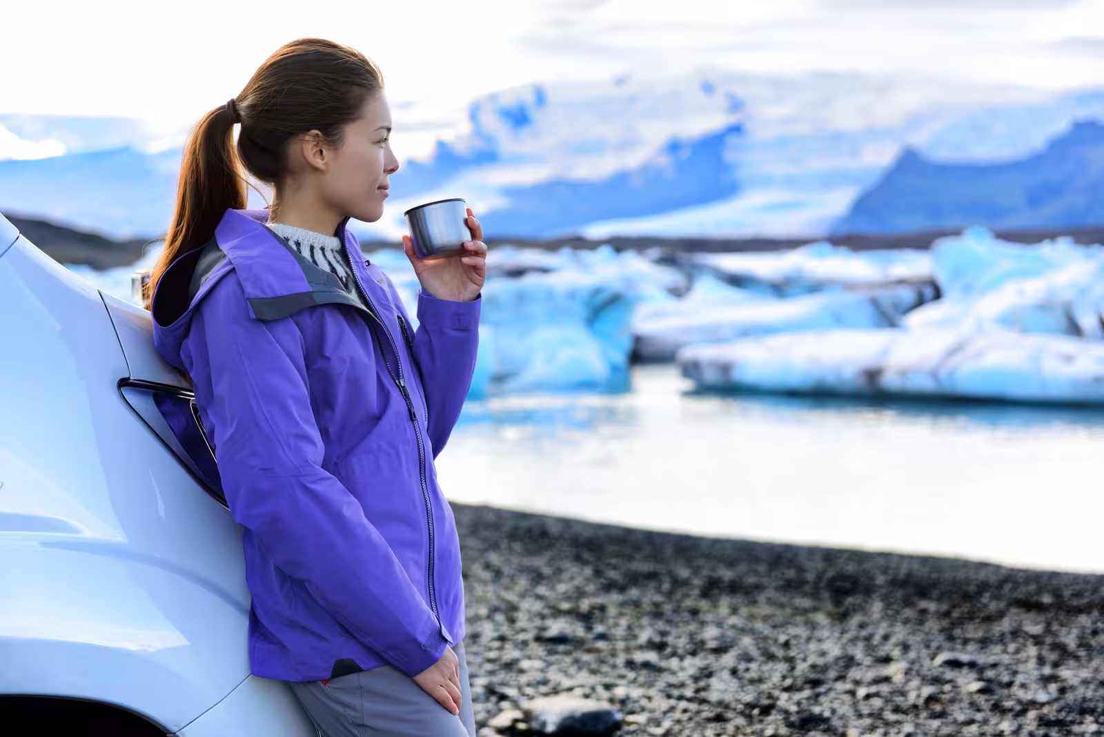 Woman in purple jacket enjoys hot drink by car with panoramic view of Icelandic glaciers and icebergs as backdrop.