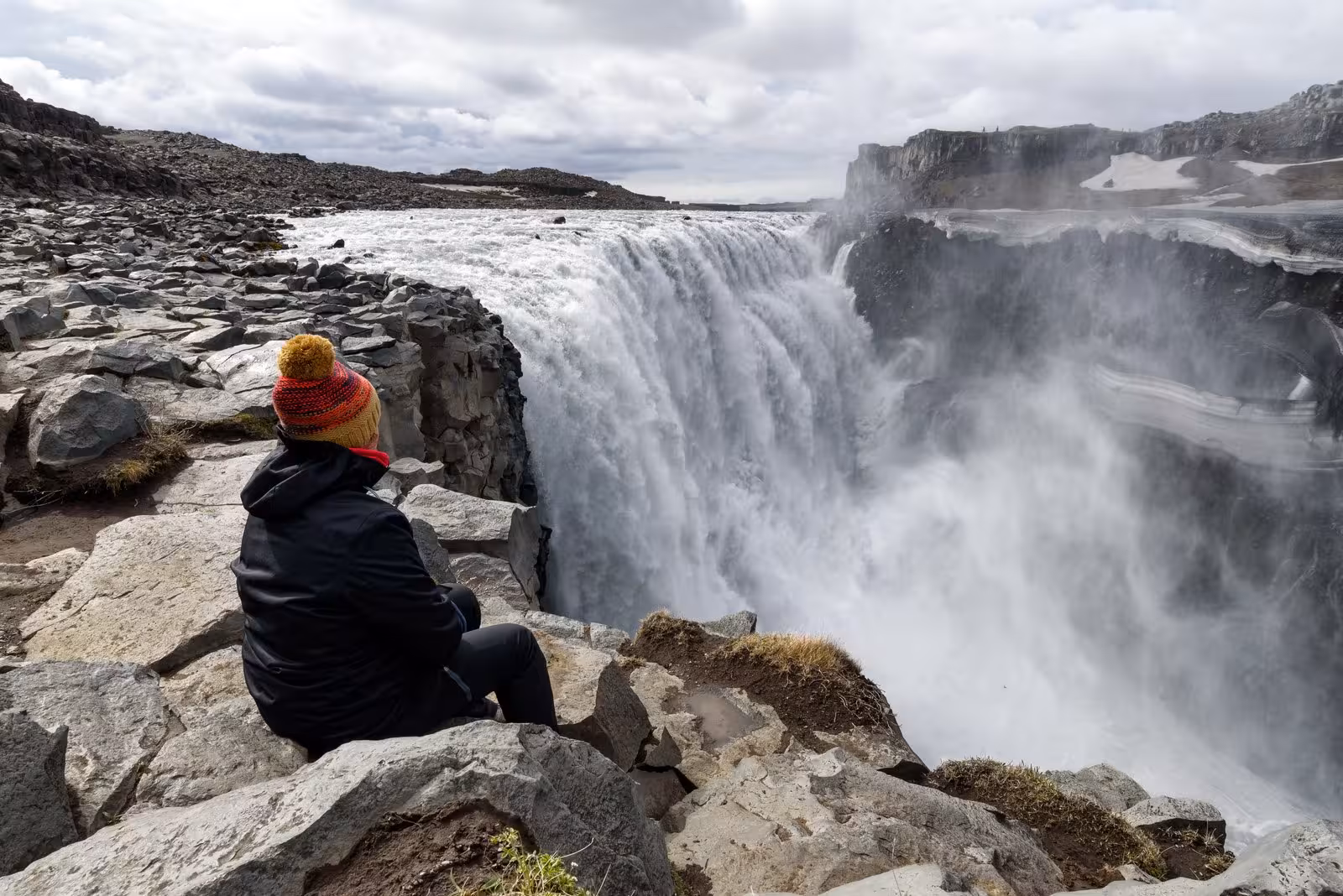 Person in warm clothing sitting by powerful waterfall, enjoying Iceland's natural beauty on self-drive road trip.