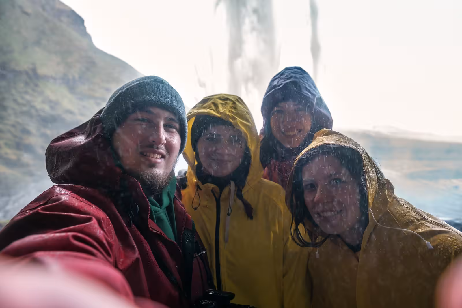 Happy travelers in rain gear taking a selfie near a waterfall, showcasing the adventure of Iceland's self-drive tour.