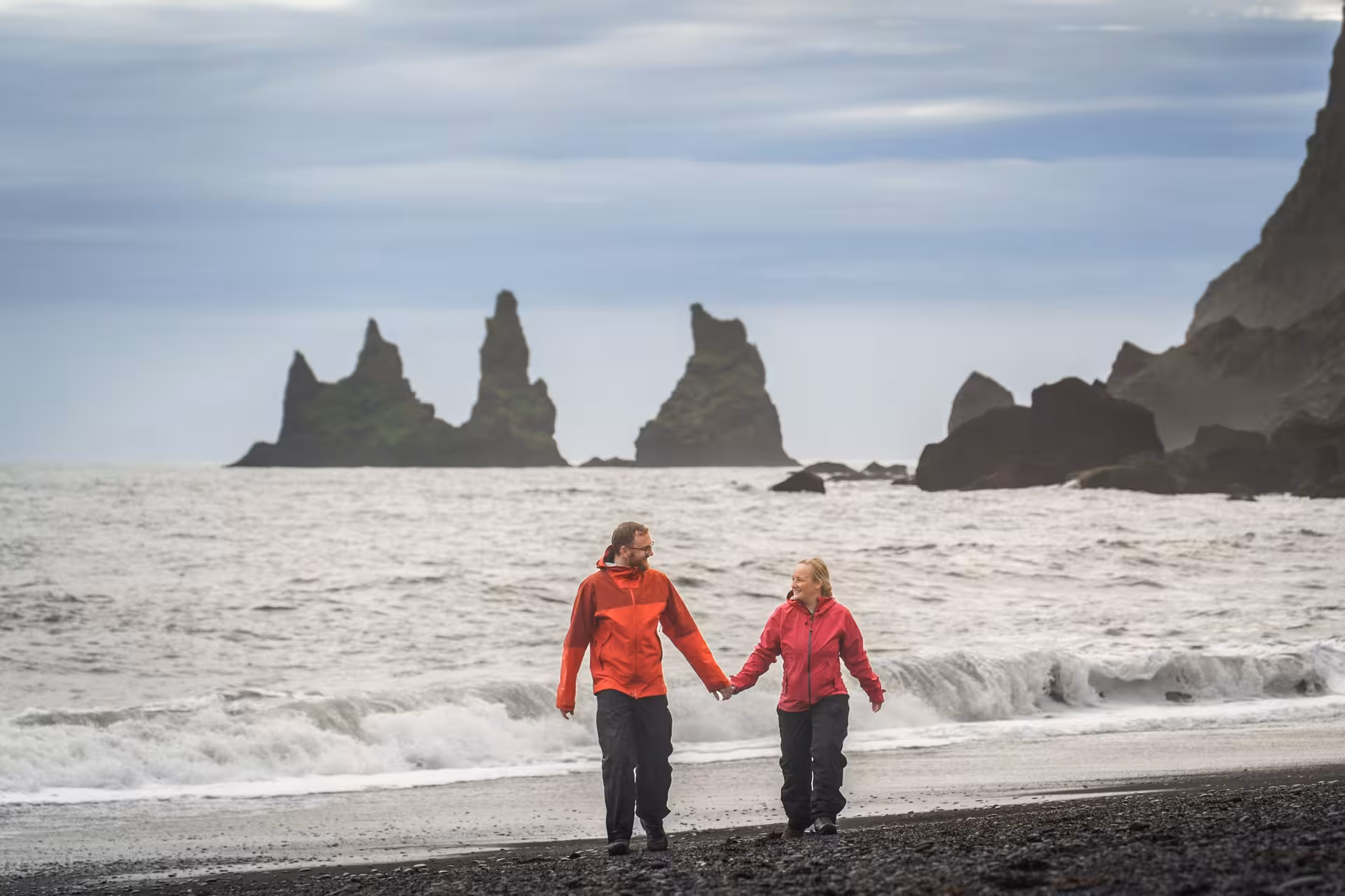 Couple exploring Reynisfjara black sand beach with dramatic sea stacks on Iceland stopover tour.