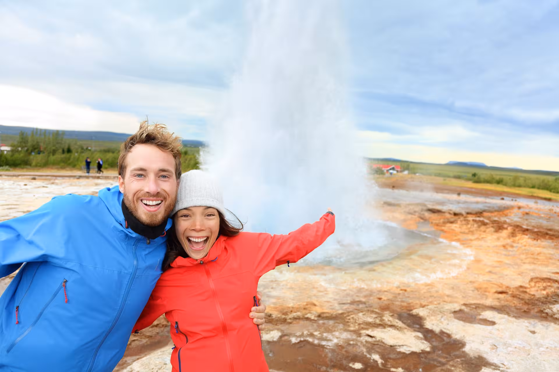Happy tourists in colorful jackets enjoying a Strokkur geyser eruption, highlighting the excitement of Iceland tours.