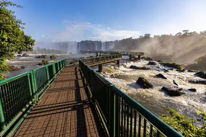 Scenic walkway view of Iguazu Falls with mist rising and sunlight illuminating the lush landscape in Brazil.