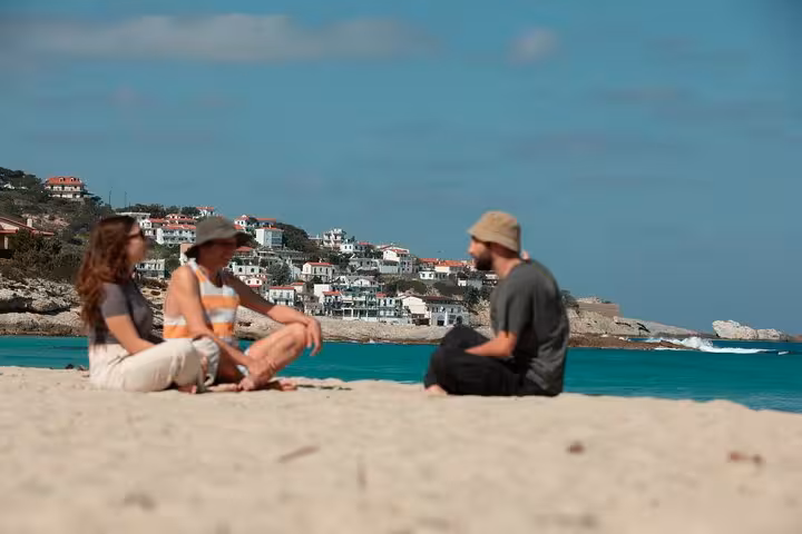 Travelers relax on a sandy Ikaria beach with turquoise Aegean Sea and traditional whitewashed village in the background