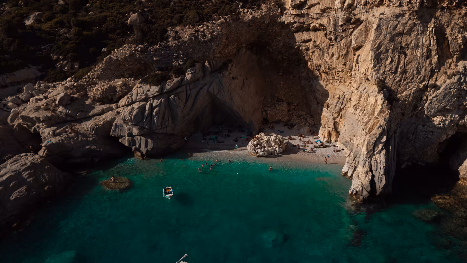 Secluded Ikaria cove with turquoise Aegean Sea, swimmers and sunbathers on a small pebble beach beneath dramatic cliffs