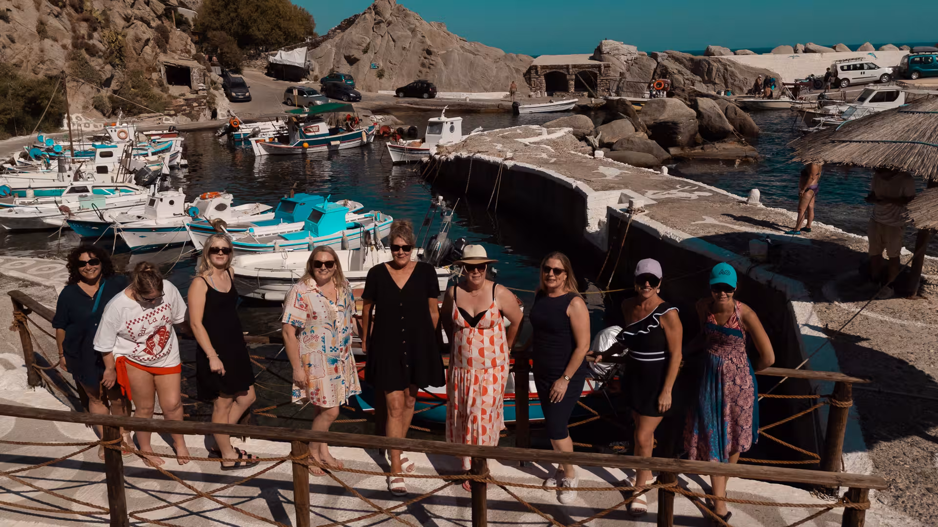 Small tour group poses by colorful fishing boats in a traditional Ikaria harbor, showcasing village charm on a guided day trip