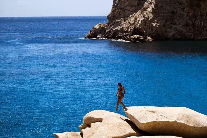 Traveler explores Ikaria’s west coast, standing on smooth granite rocks above crystal-clear Aegean Sea waters