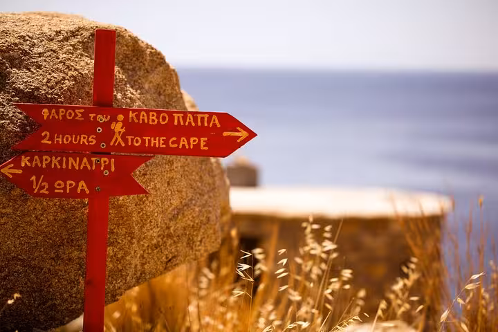 Red hiking trail sign in Greek pointing to Ikaria west coast landmarks, with golden grass and Aegean Sea in the background