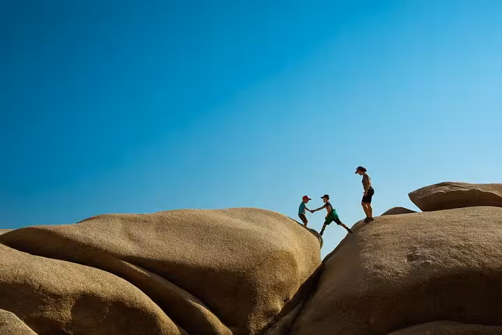 Friends hiking on smooth granite rocks in Ikaria, helping each other climb under a clear blue sky on the island’s wild west coast