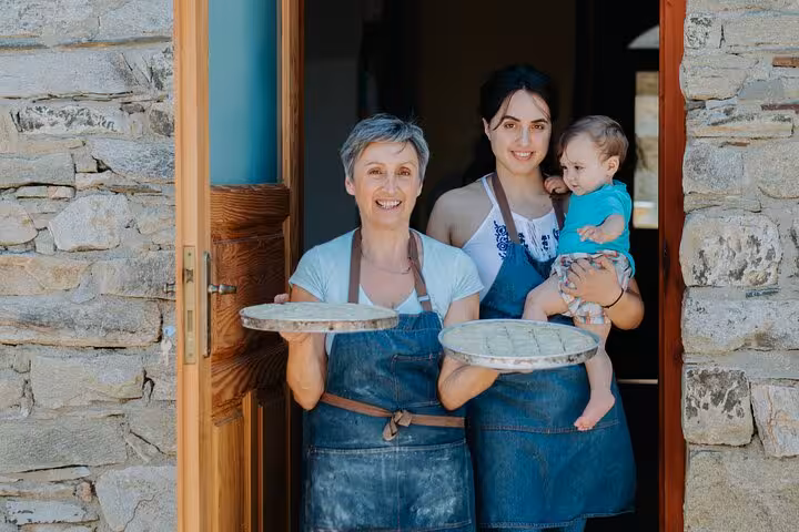 Smiling Ikarian women in aprons holding homemade pies at a stone farmhouse doorway on the Ikaria Longevity family winery tour