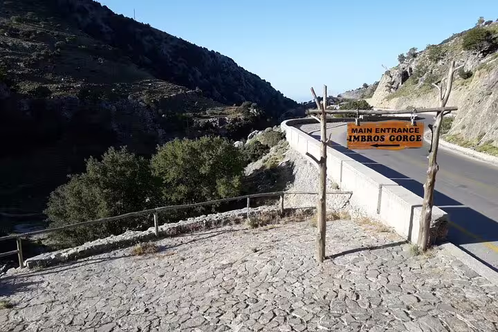 Entrance to Imbros Gorge marked by a wooden sign, set against a backdrop of rocky hills and clear blue skies.