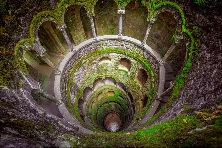 Enchanting view of the Initiation Well at Quinta da Regaleira in Sintra, showcasing moss-covered spiral stairs and arches.