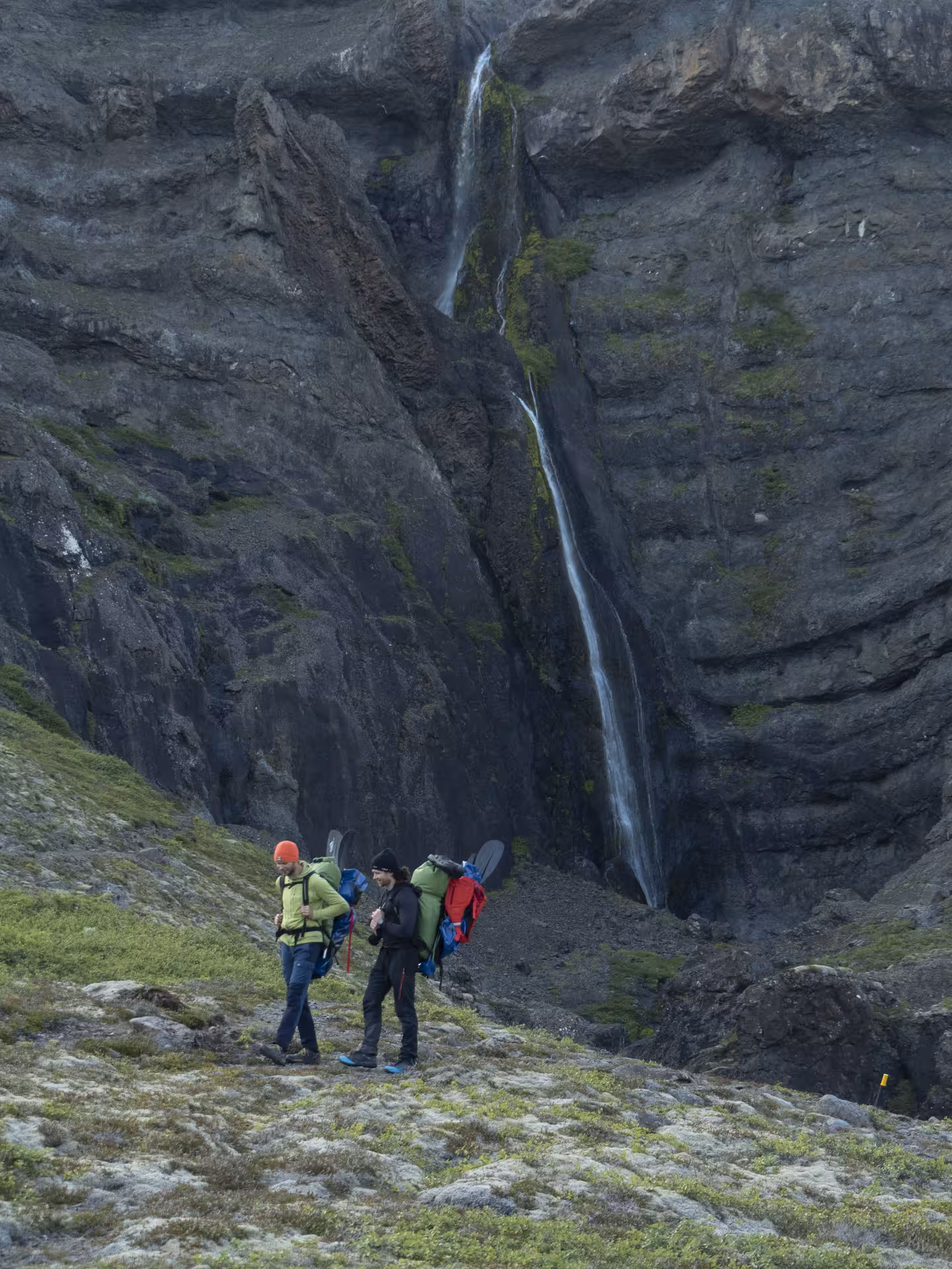 Hikers carry packrafts and gear past a waterfall on a glacier valley approach for intro packrafting tour
