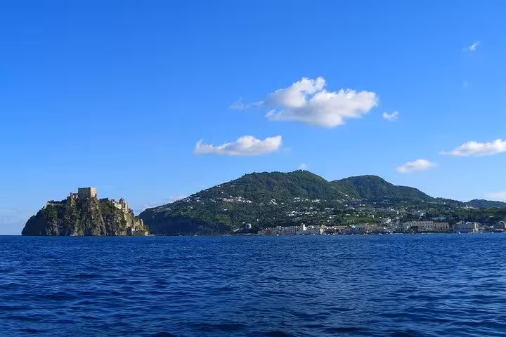 Picturesque view of Ischia's Aragonese Castle against a backdrop of lush green hills and clear blue sea on a sunny day.