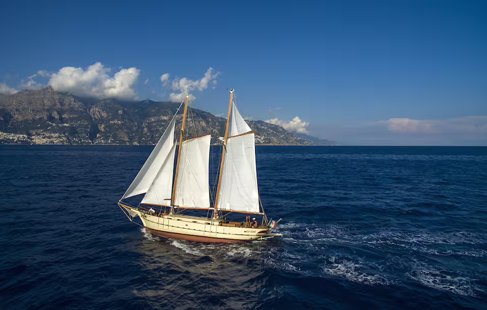 Classic sailboat cruising off the coast of Ischia, Italy, with blue Tyrrhenian Sea and rugged cliffs in the background