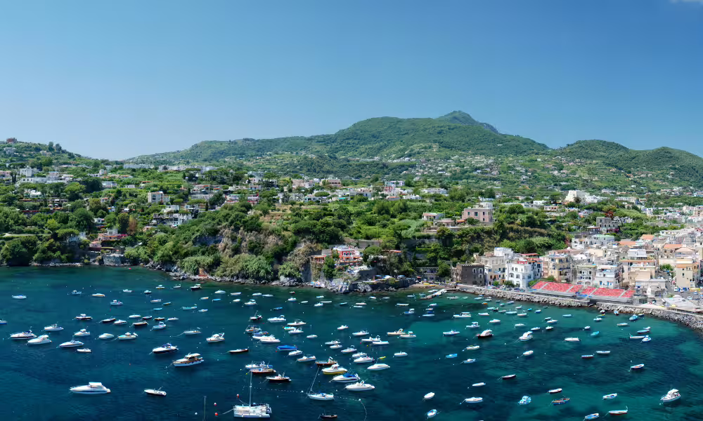 Boats anchored in a turquoise bay below lush green hills and pastel villages on an Ischia island boat excursion