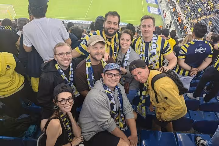 Travelers with local host at Istanbul football match, wearing scarves in stadium stands before kickoff