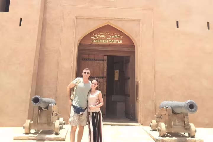 Visitors pose by the entrance of historic Jabrin Castle in Nizwa, Oman, highlighting traditional architecture and cannons.