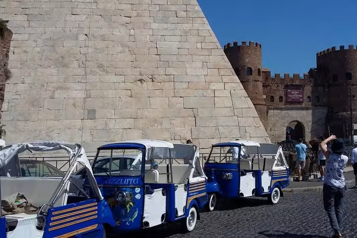 Line of blue Ape Calessinos parked near historic Roman architecture during a James Bond tour.