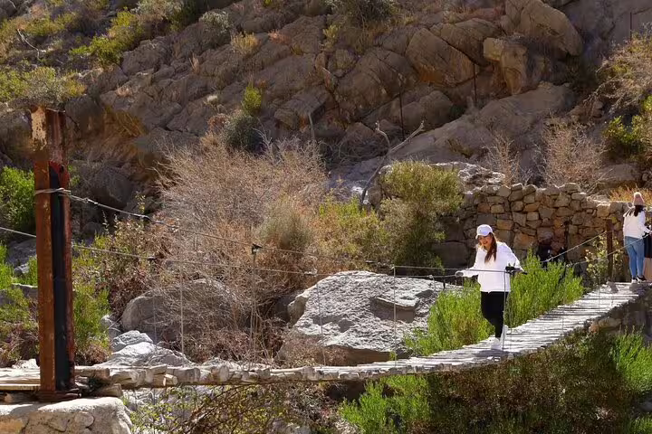 Tourists crossing a rustic bridge surrounded by rocky terrain in Jebel Akhdar Al Suwjara Village, Oman.