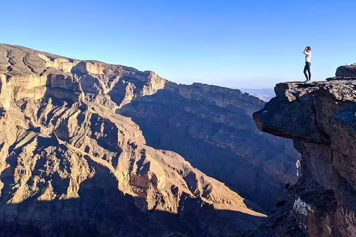 Person stands at the edge of Jebel Shams cliff, admiring expansive canyon views in bright sunshine on a guided hike.