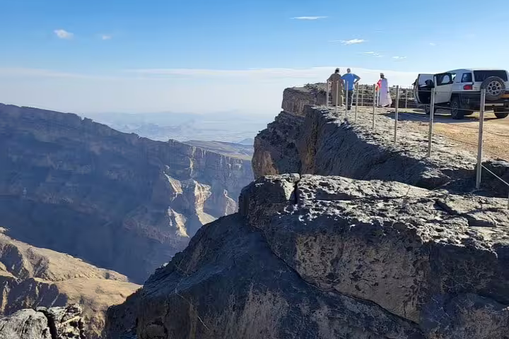 Tourists and a vehicle on a rocky path enjoying panoramic views of Jebel Shams canyon on a private guided hiking tour.