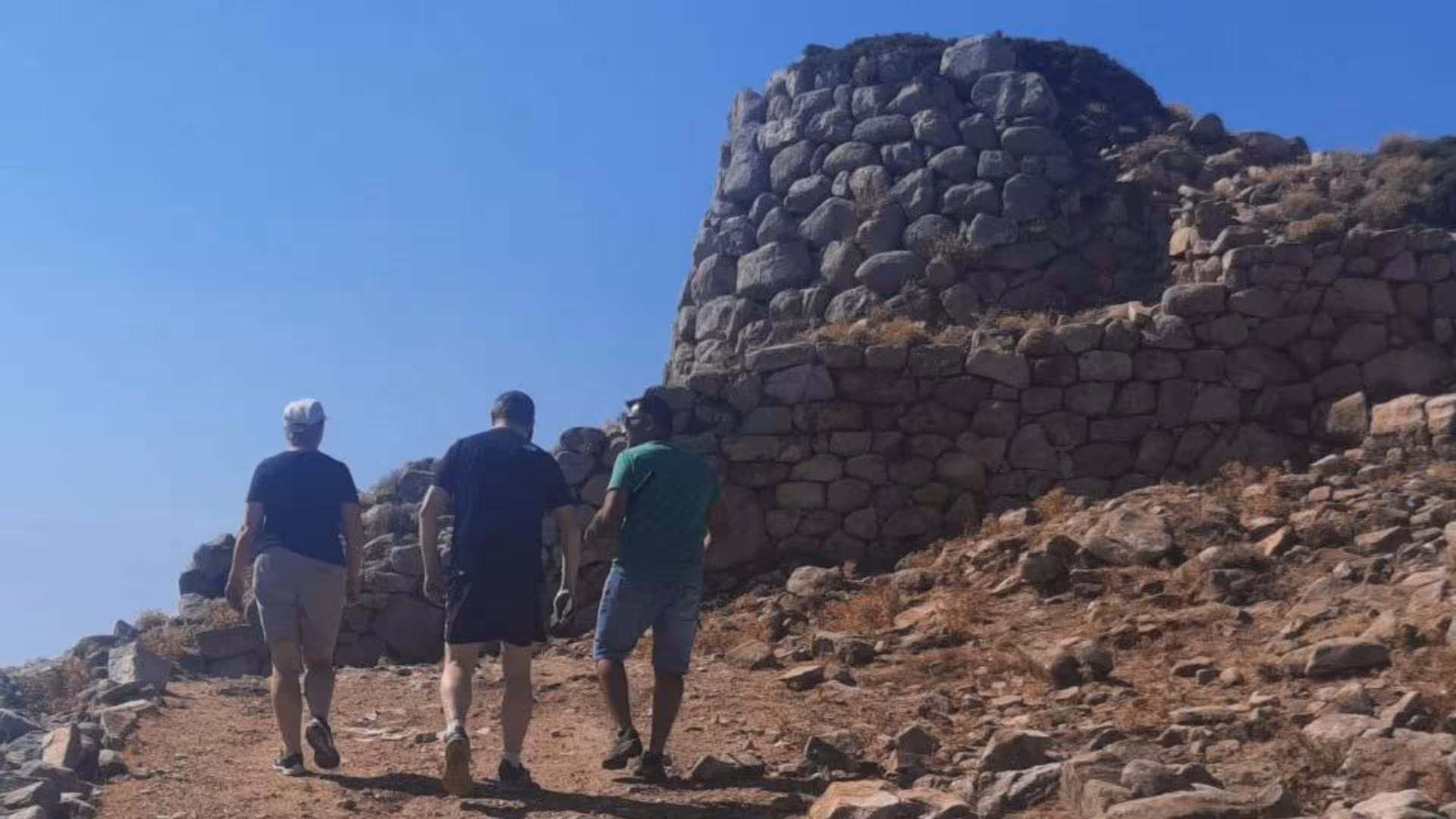 Three tourists exploring ancient stone ruins on a rugged hillside under a clear blue sky in Barbagia, Sardinia.