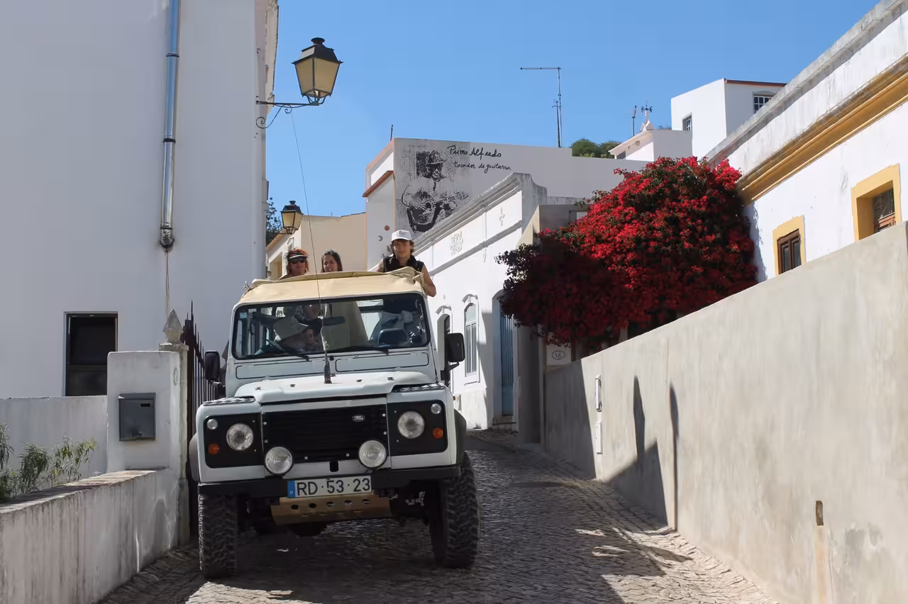 Open-top 4x4 Jeep Zoofari drives through a narrow Algarve village street with white houses and flowers