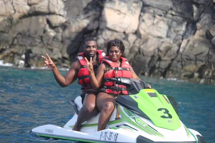 Joyful duo on a jet ski, making peace signs during a coastal safari adventure with rocky cliffs in the background.