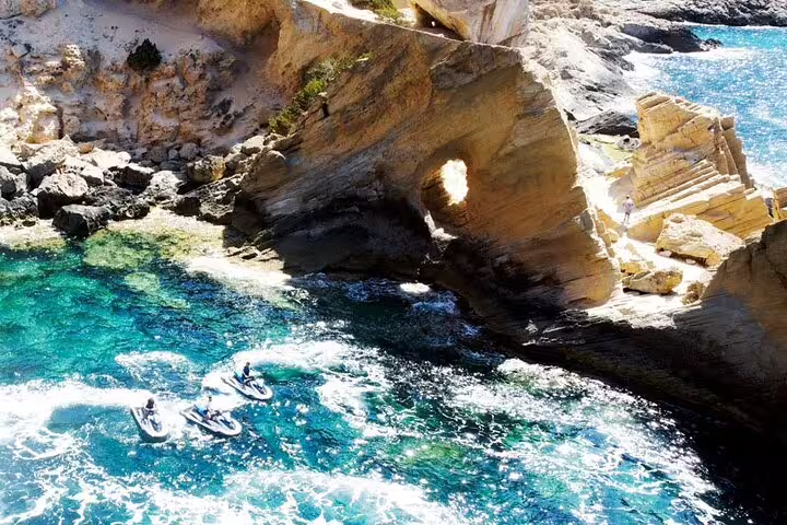 Jet ski tour from San Antonio Ibiza to Atlantis, riders cruising turquoise cove by dramatic sea arch cliffs