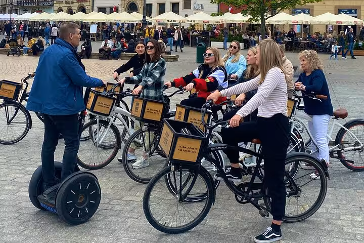 A group of cyclists listening to a guide in the Jewish Quarter during a bike tour, surrounded by a lively plaza.